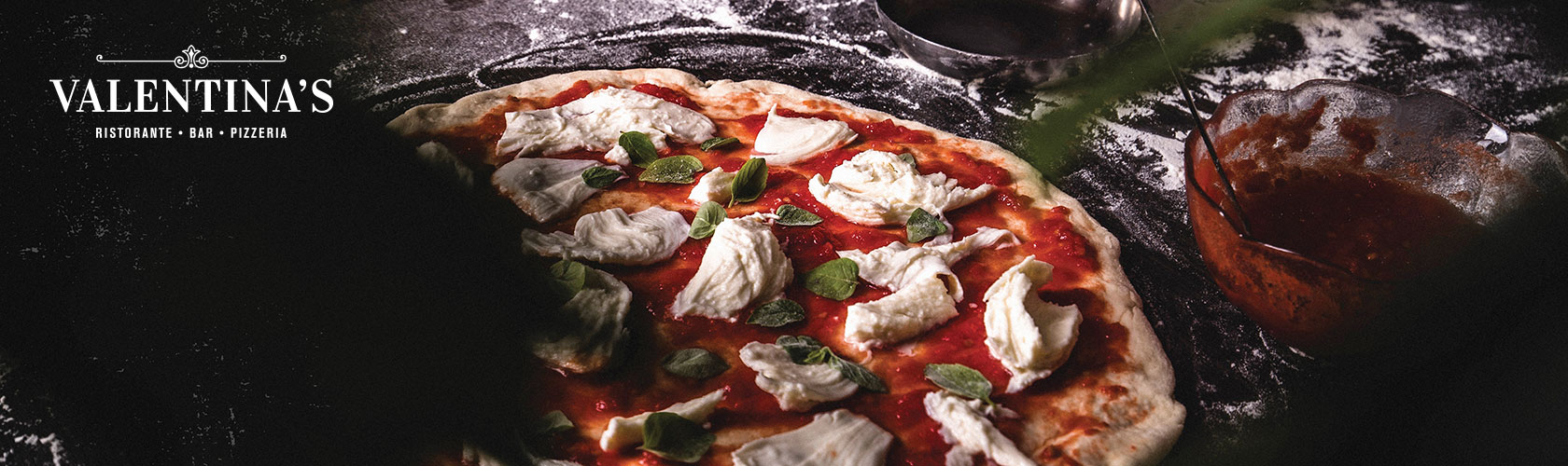 Artisan pizza with small bowl of red sauce next to it and Valentina's logo in upper left corner.